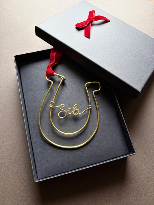 A gold wire horseshoe ornament with the name 'Josh' written in cursive, presented in an open black gift box with a red ribbon on a brown background.