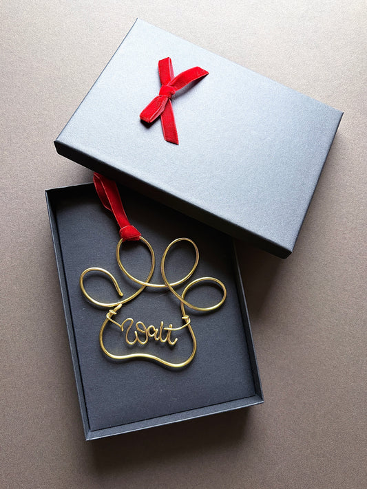 A gold wire paw-shaped ornament with the word 'Walt' in script, displayed inside an open dark gray gift box with a red ribbon. The lid of the box is placed above, showing a red bow, on a neutral brown background.