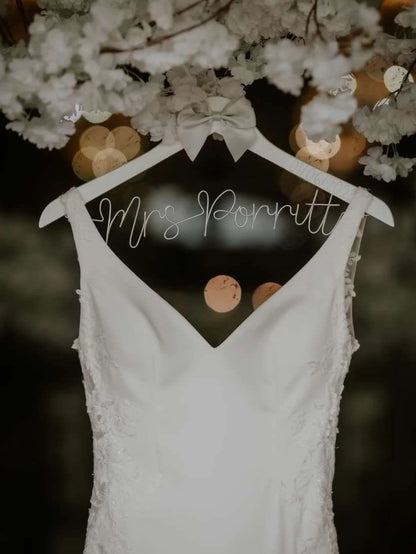 white wedding dress hung on a personalised hanger, with silver wire lettering and silver satin bow
